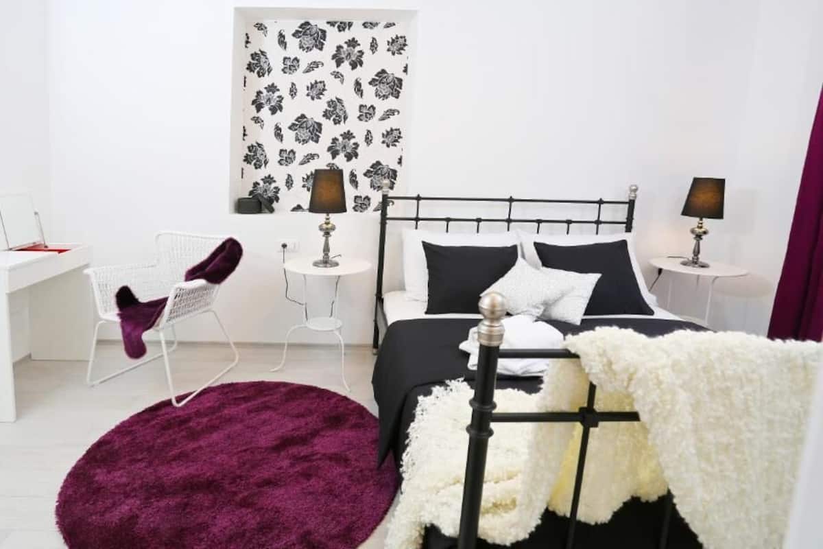 Un dormitorio con estructura de cama metálica negra, ropa de cama blanca, un tapete morado, una silla blanca y una mesita de noche con lámpara.