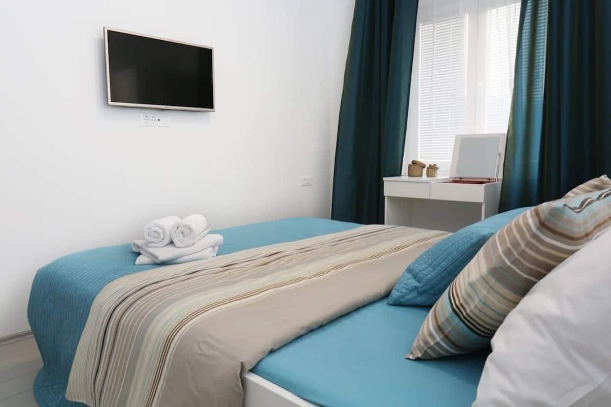 Un dormitorio con cama, televisor montado en la pared, escritorio con ordenador portátil y ventana con cortinas.