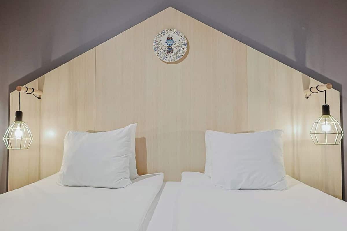 Habitación de hotel con cabecera de madera, dos camas con ropa blanca y lámparas fijadas a la pared.
