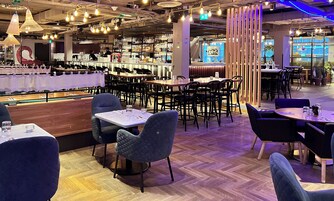 Interior de un restaurante con varias mesas, sillas y una zona de bar con estantes llenos de botellas.