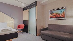 Habitación de hotel con una cama, un sofá gris, una silla roja, una puerta de cristal y un cartel en la pared.