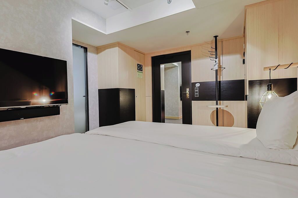 Ein Hotelzimmer mit flachbildfernseher an der Wand, einem Bett mit weißer Bettwäsche, einem Holzschrank und einer modernen Leuchte.