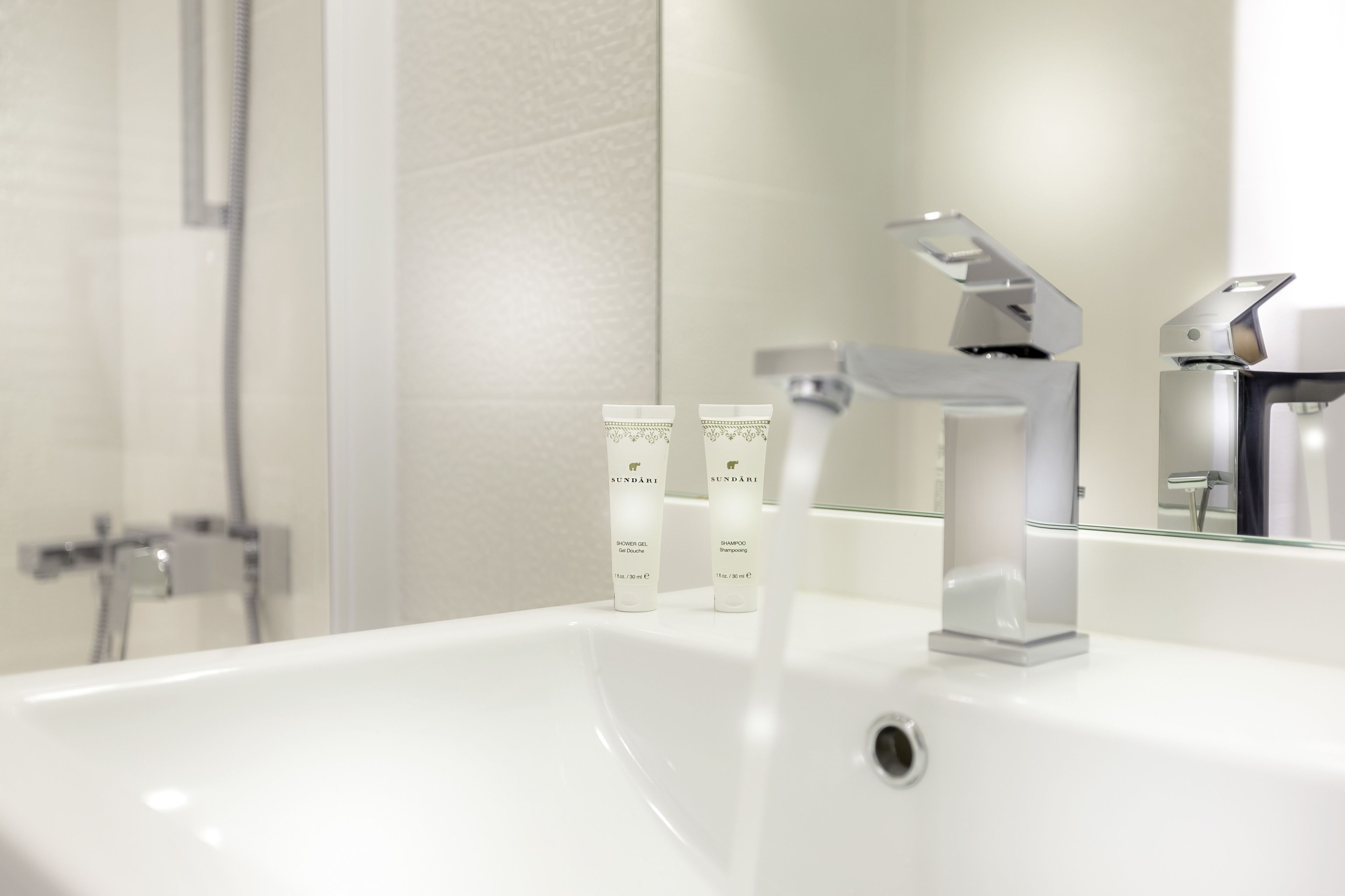 A modern bathroom with a white sink, a handheld showerhead, and two bottles of Sundari shower gel and shampoo.