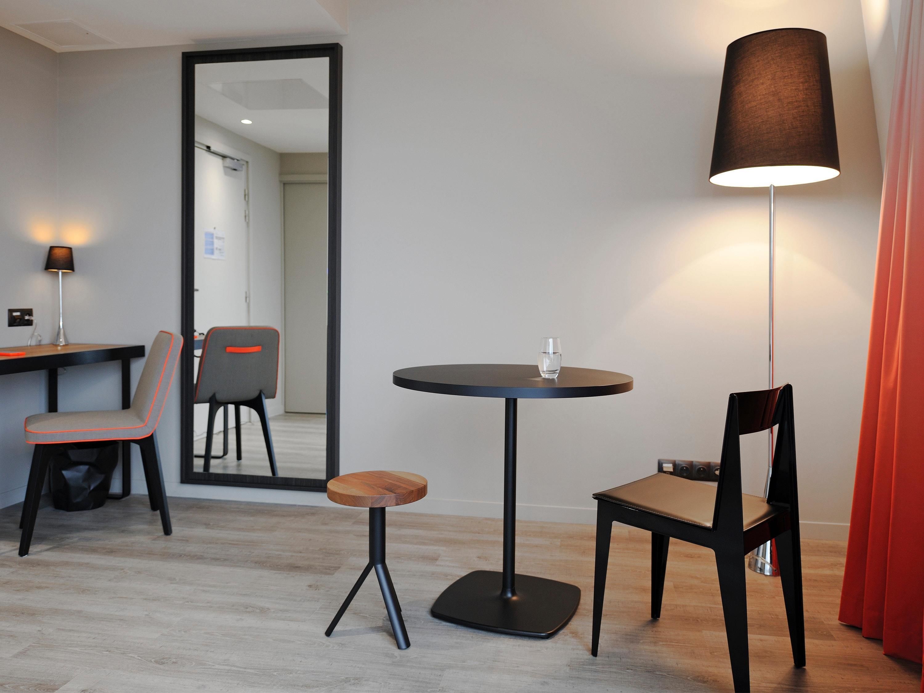 Ein modernes Hotelzimmer mit einem runden Tisch, zwei Stühlen, einem kleinen Hocker, einer Stehlampe und einem Spiegel.
