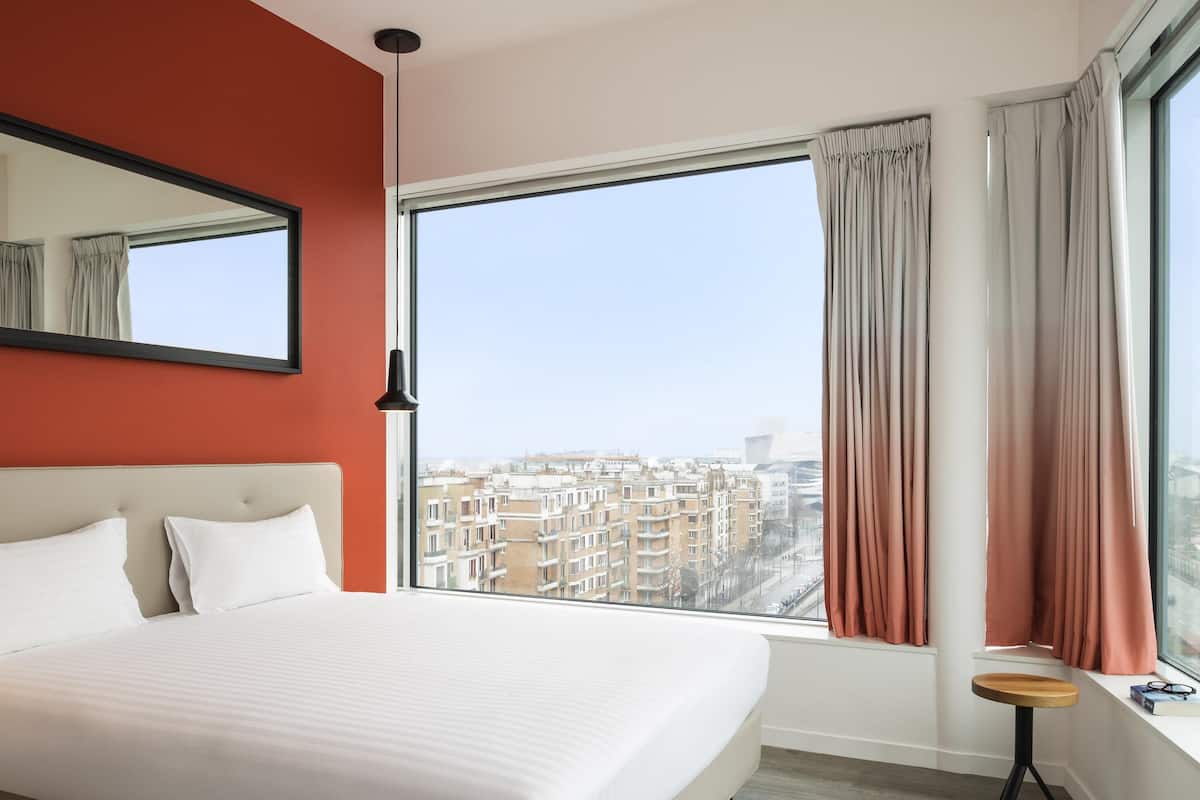 Habitació d'hotel amb un llit gran, una finestra amb cortines, una taula petita i vista a la ciutat.