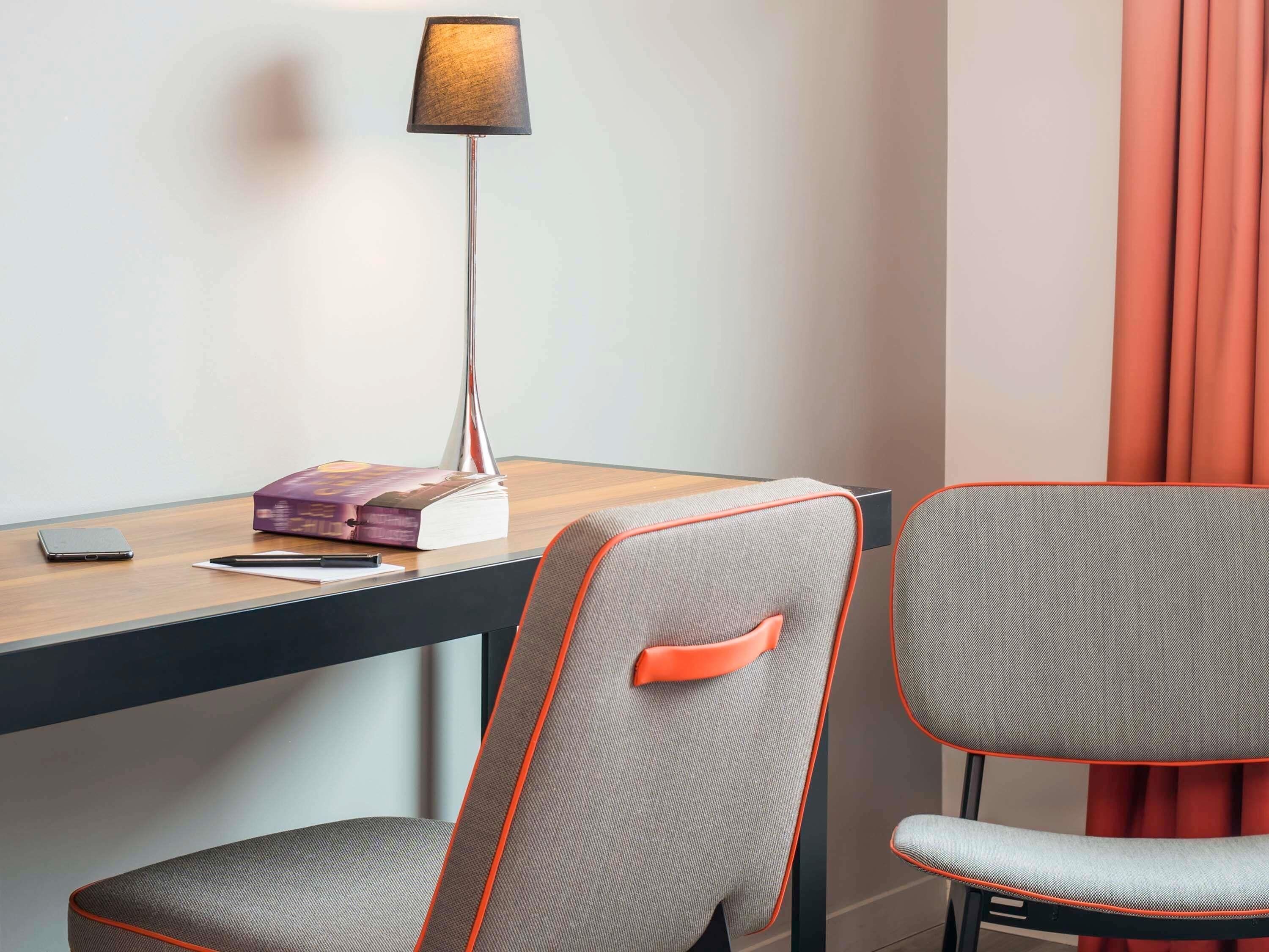 Et hotelværelse med et skrivebord, en lampe, en stol og en bog.