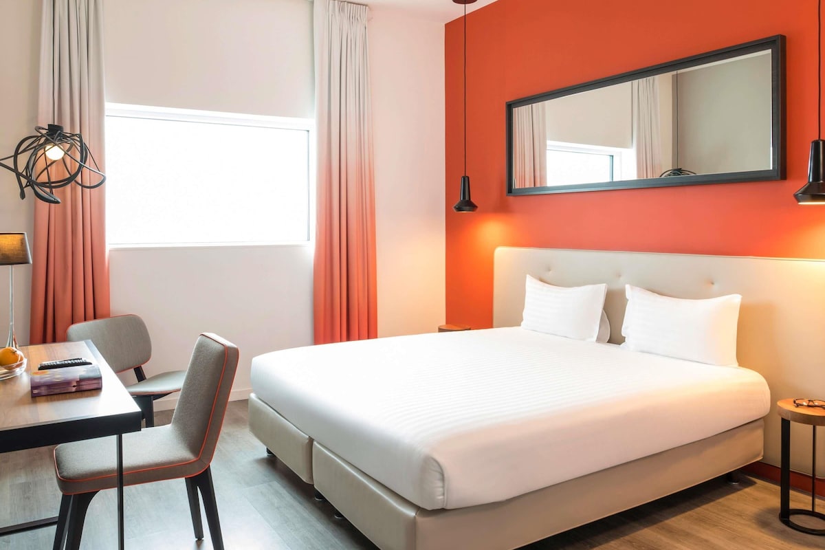 A hotel room with a bed, a desk with a chair, a small table, a lamp, and a window with curtains.