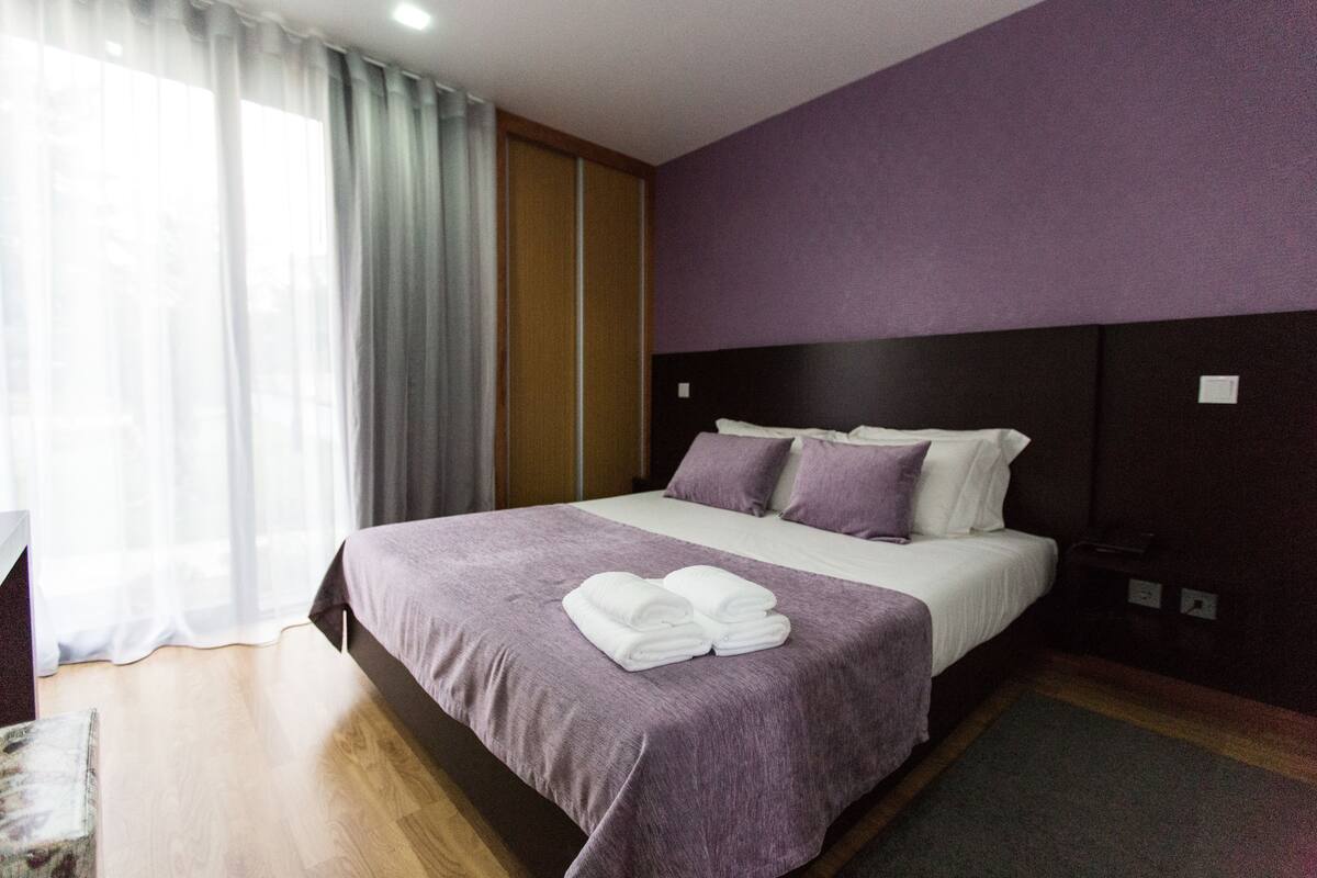 A hotel room with a bed, a headboard, a window with curtains, and a bedside table.