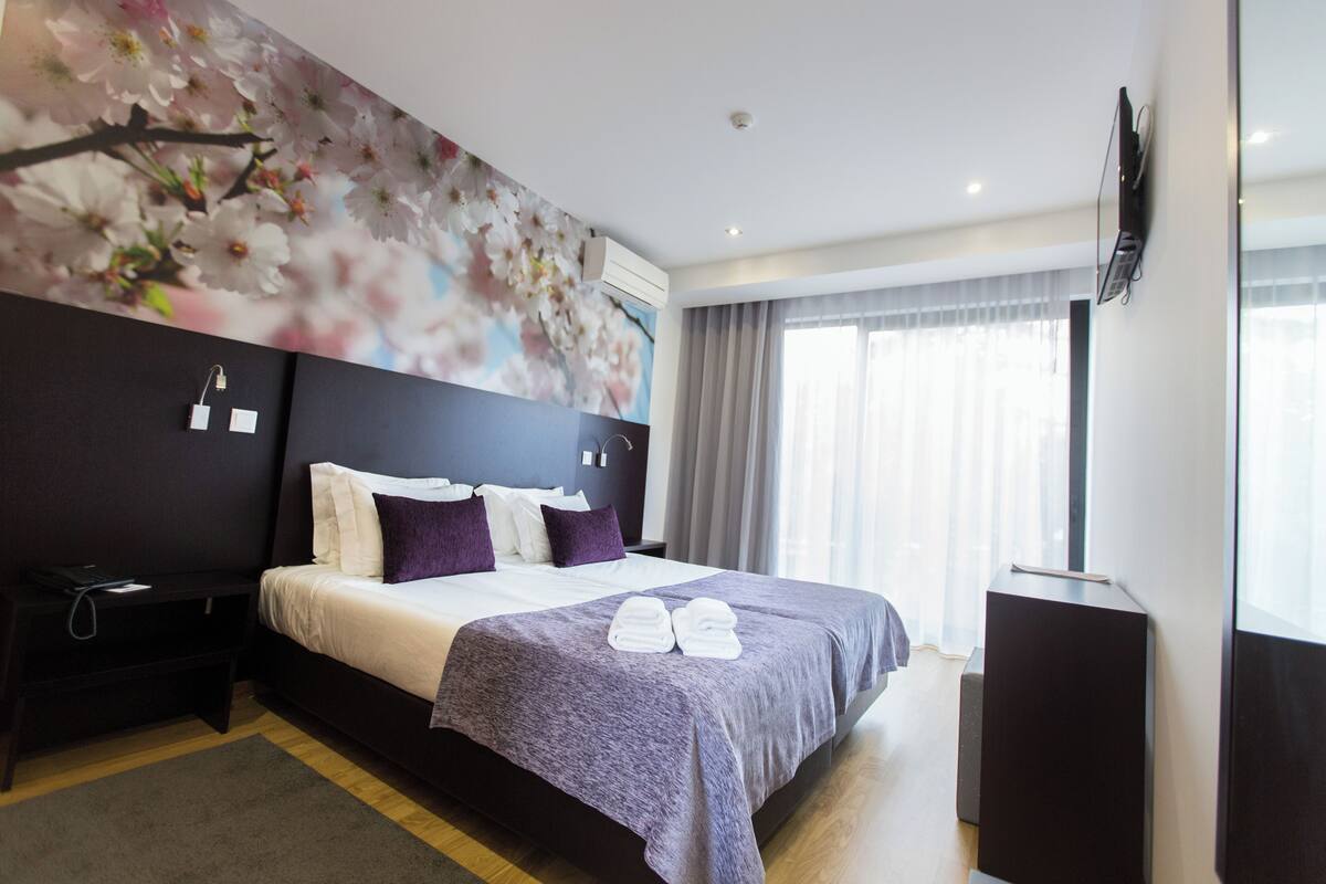 A modern hotel room with a large bed, a flat-screen TV mounted on the wall, and a window with curtains.