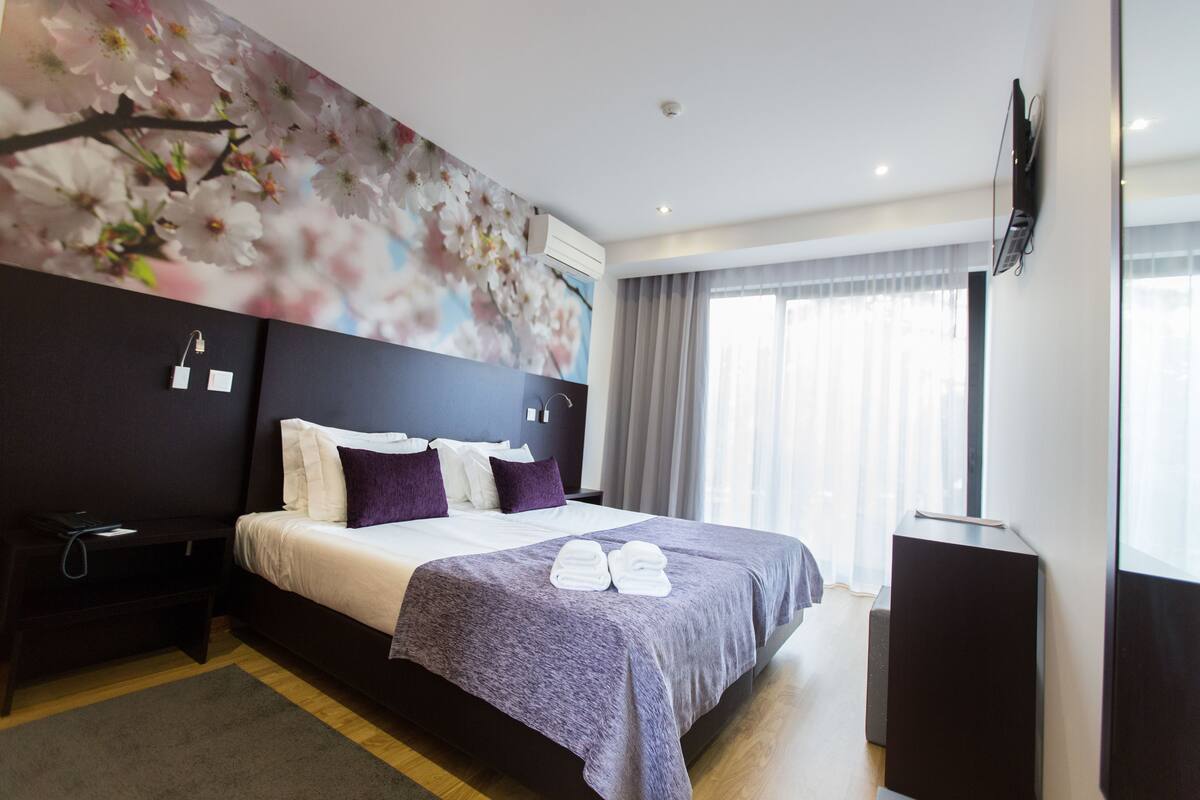 A modern hotel room with a large bed, a flat-screen TV mounted on the wall, and a window with curtains.