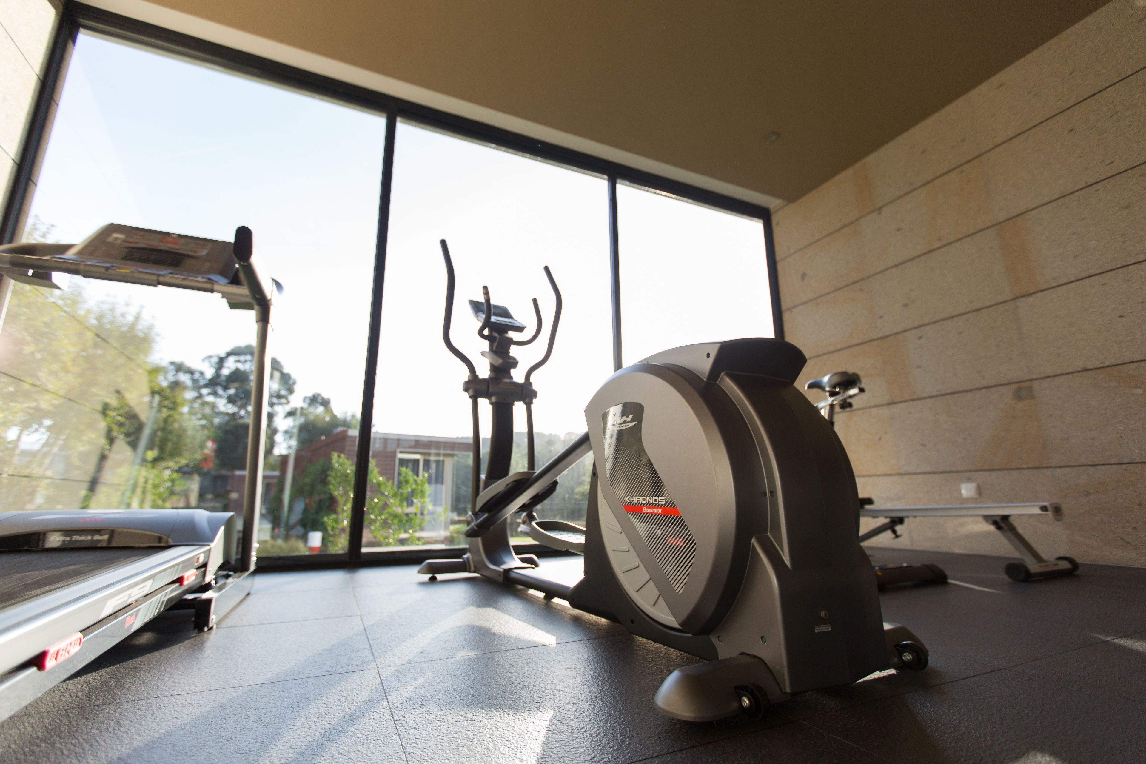 Un gimnasio con equipamiento de ejercicio, ventanas amplias y vistas a árboles y edificios.