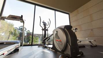 Un gimnasio con equipamiento de ejercicio, ventanas amplias y vistas a árboles y edificios.
