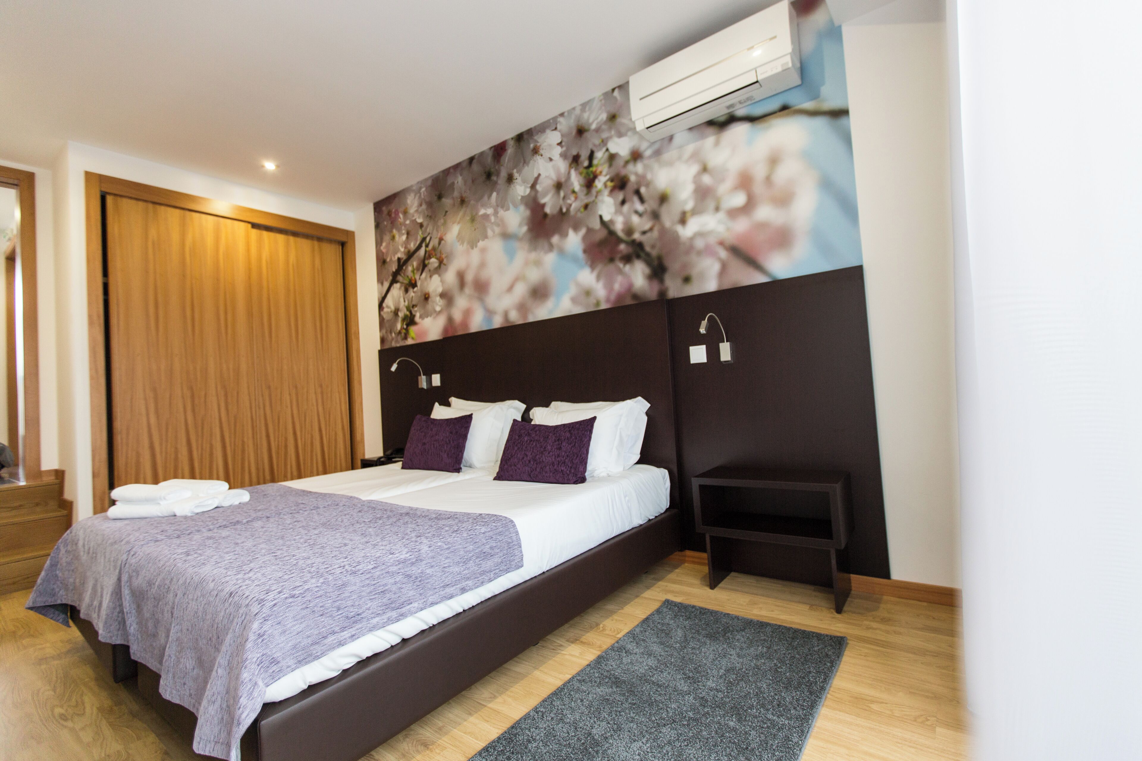 Une chambre moderne avec un grand lit, une armoire en bois, une petite table de chevet et un climatiseur fixé au mur.
