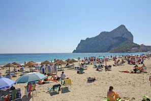 A crowded beach with numerous sun loungers and umbrellas, a prominent rocky cliff in the background, and a clear blue sky. - Coral Beach (Calpe)