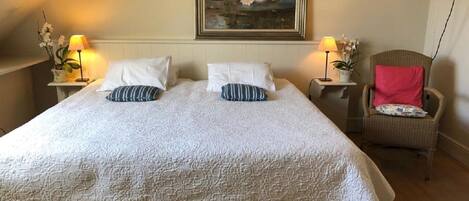 A bedroom with a large bed, two bedside tables with lamps, a chair with a pink cushion, and a painting on the wall.