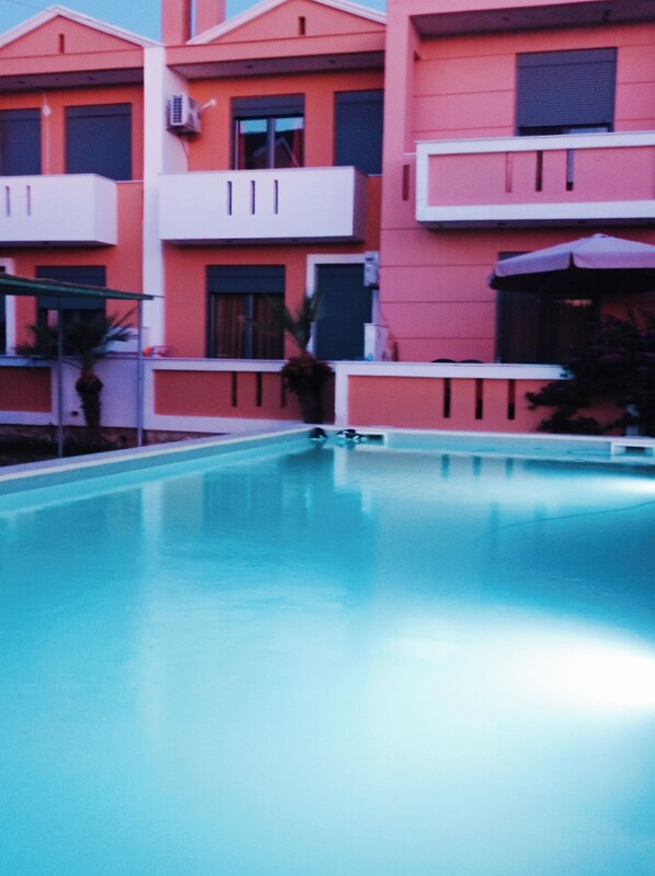 A swimming pool with a clear blue water surface, surrounded by a building with a pink facade and white balconies. - Nitelli's Houses (Lesvos)