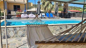 Una hamaca con vista a una piscina con sillas de descanso y sombrillas, frente a un muro de piedra y un edificio con tejado rojo.