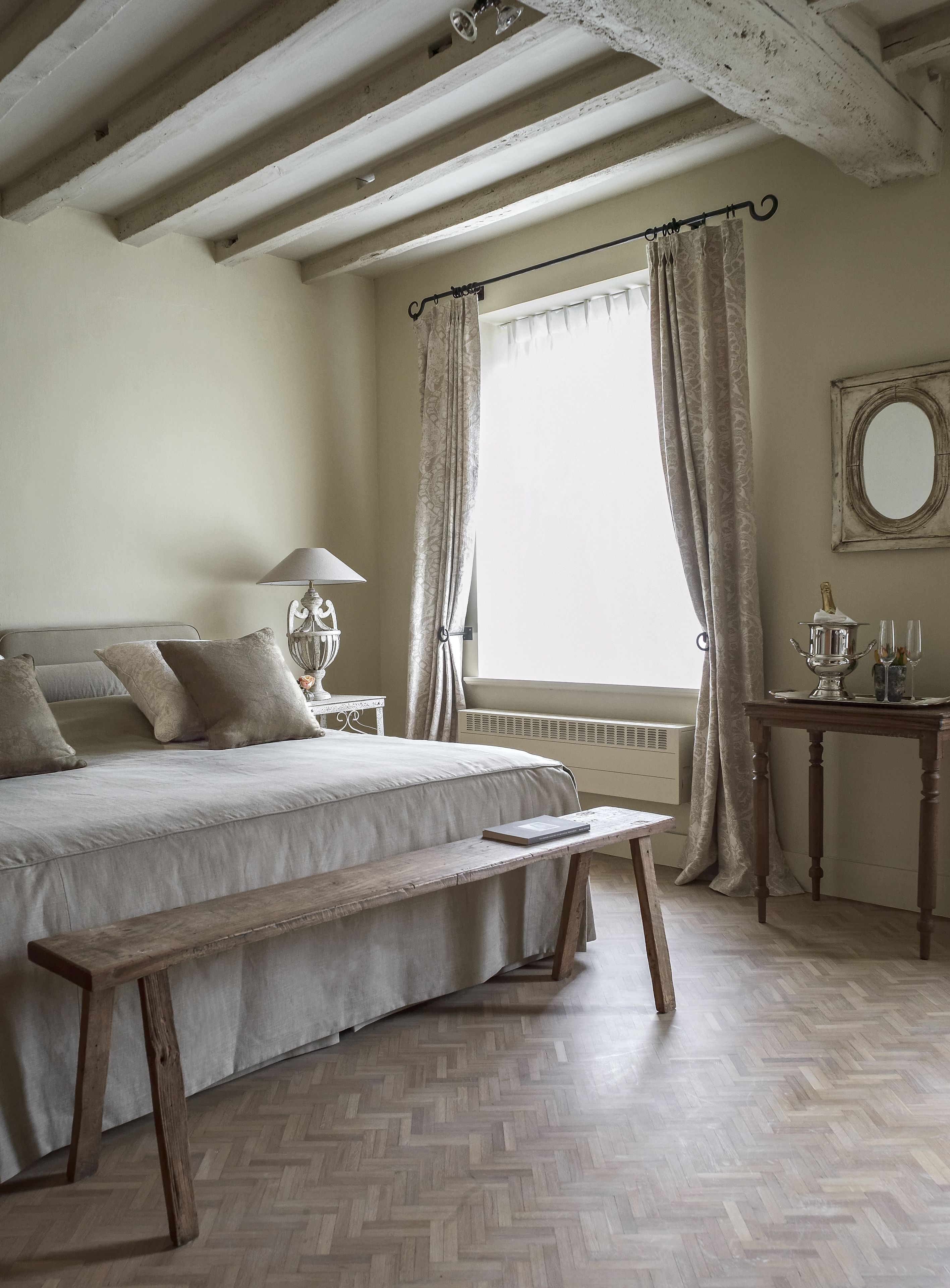 A bedroom with a bed, a bedside table with a lamp, a bench, a window with curtains, and a mirror on the wall.