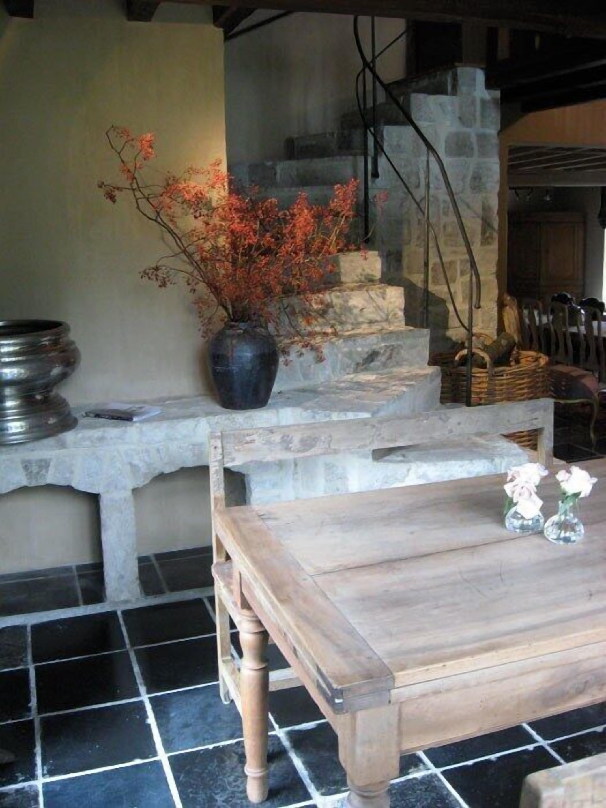 Un interior rústico con un banco de piedra, una mesa de madera y un jarrón con flores rojas.