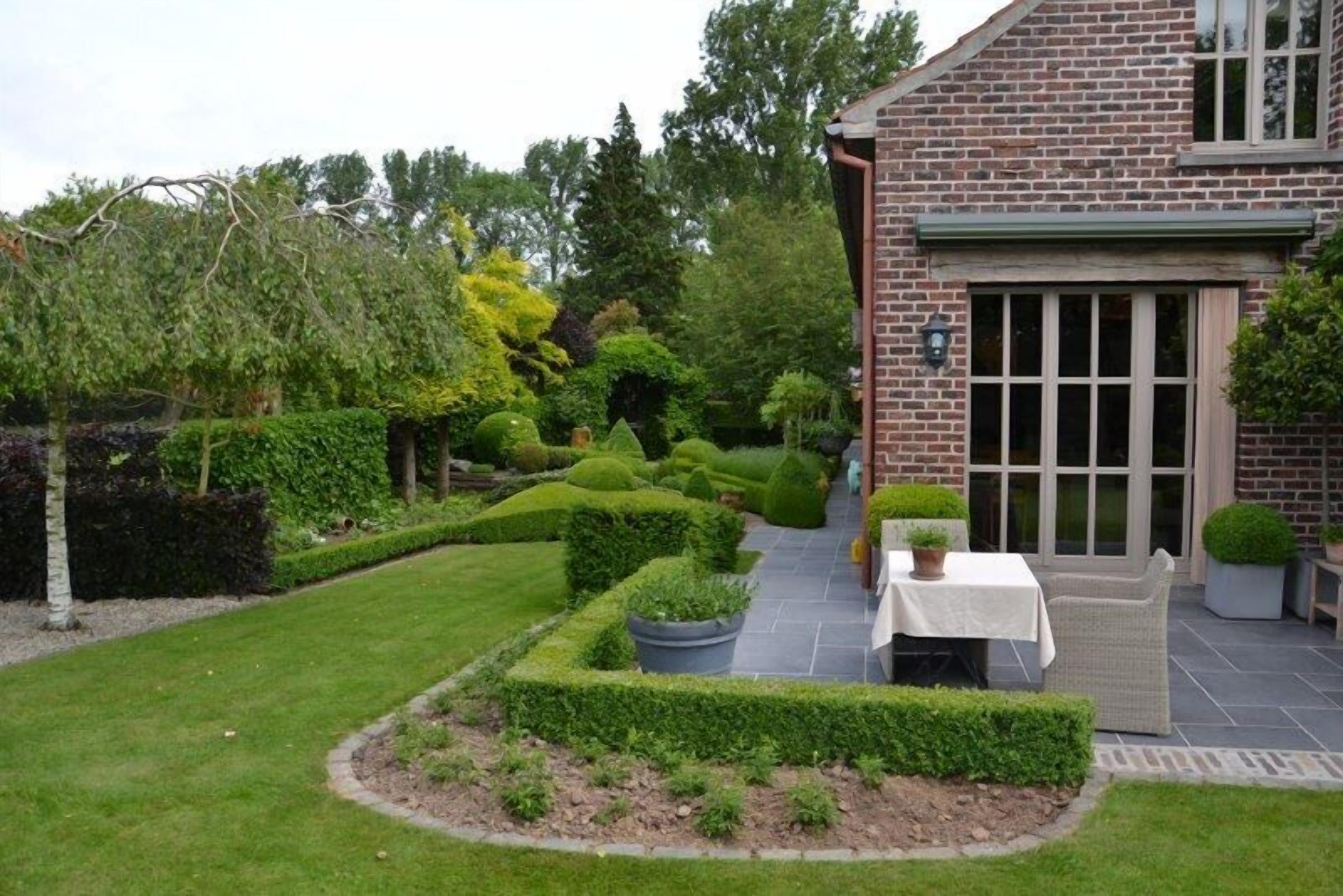 A well-maintained garden with a brick building, a patio table, and neatly trimmed hedges.