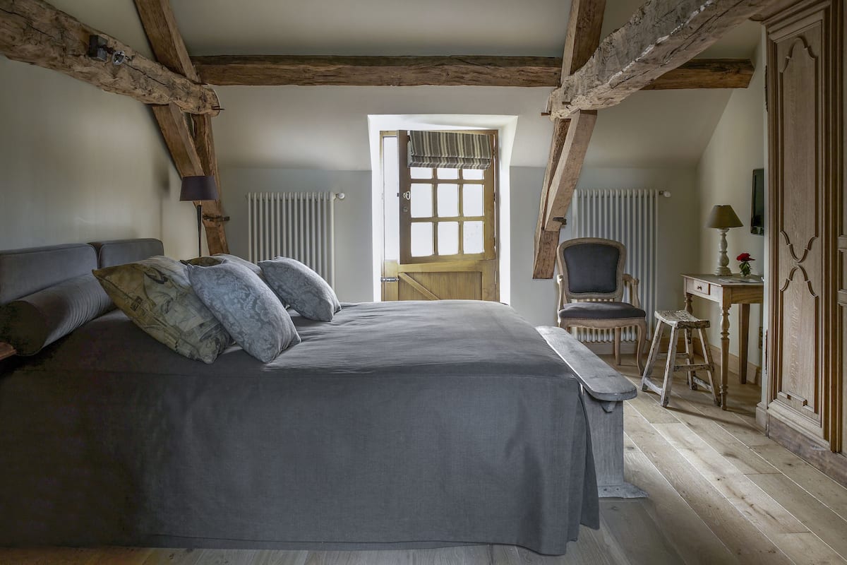 A bedroom with a large bed, wooden beams, a radiator, a chair, and a desk with a lamp.