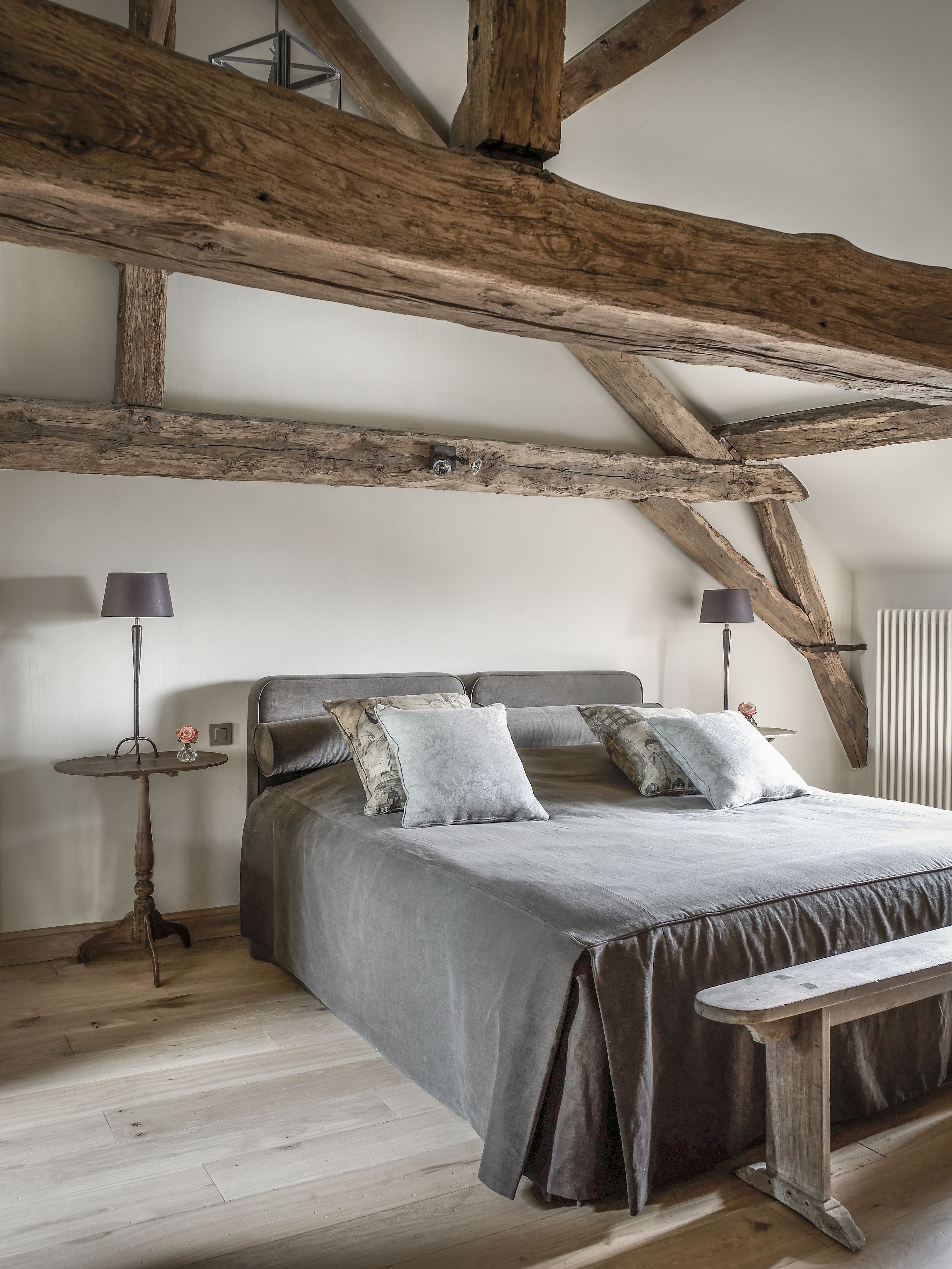 A bedroom with a large bed, wooden beams, a small table with a lamp, and a bench.