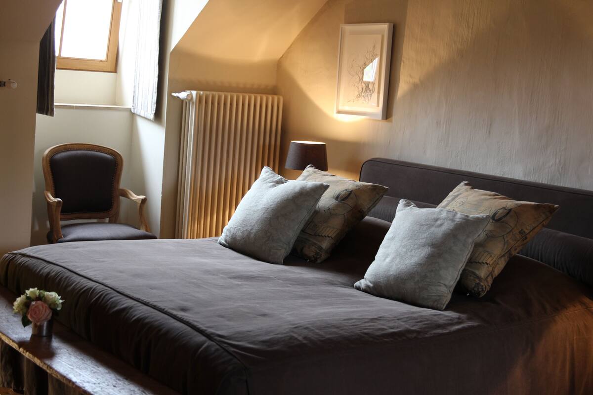 A bedroom with a large bed, a chair, a bedside table, a lamp, a radiator, and a framed picture on the wall.