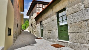 A narrow cobblestone street flanked by stone buildings with green doors and windows, leading to a hill in the distance. - Aldeia do Tâmega (Amarante)