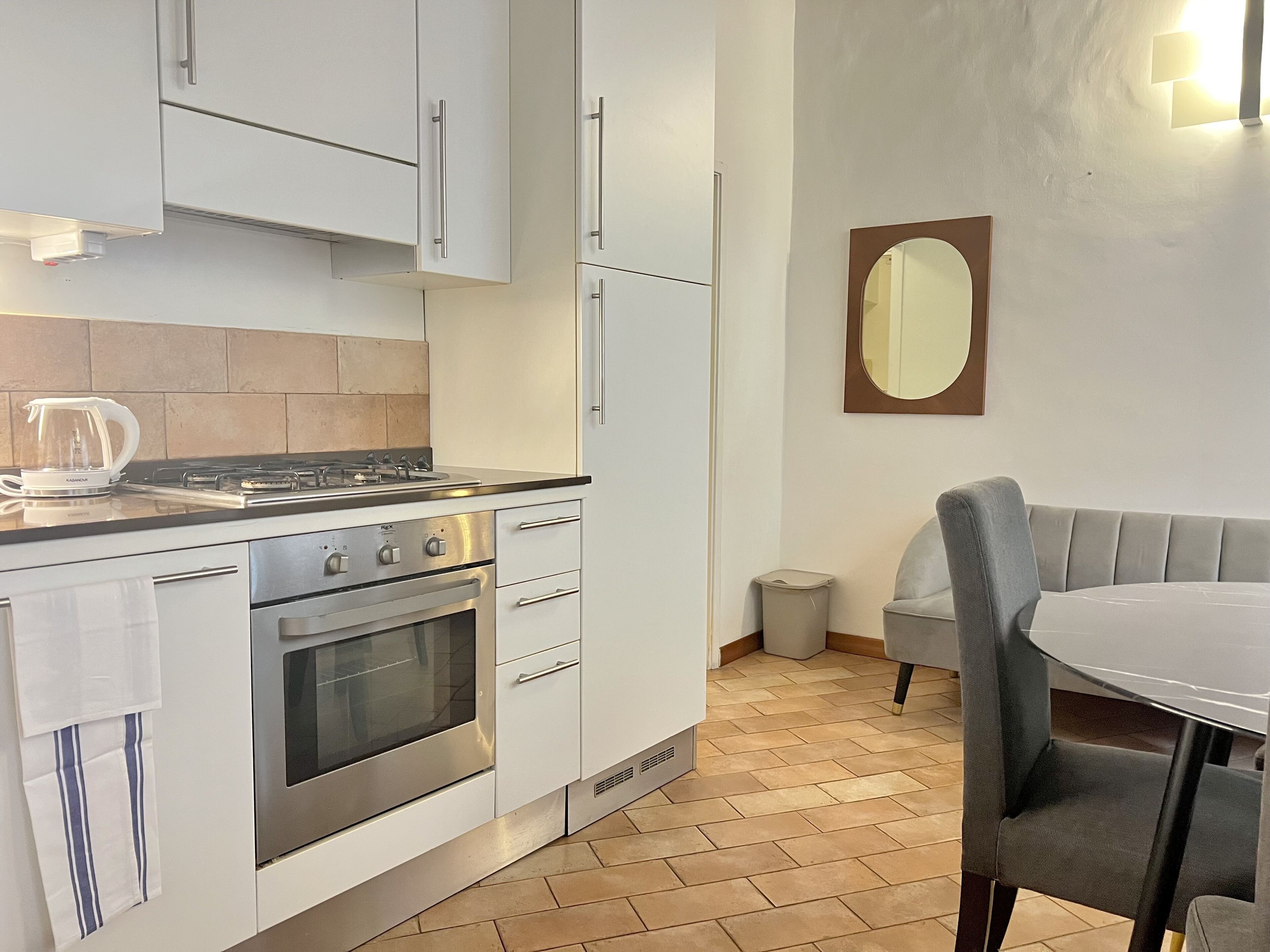 Una cucina moderna con mobili bianchi, un fornello a gas e un forno. Si nota un cestino e una sedia grigia vicino a un tavolo.