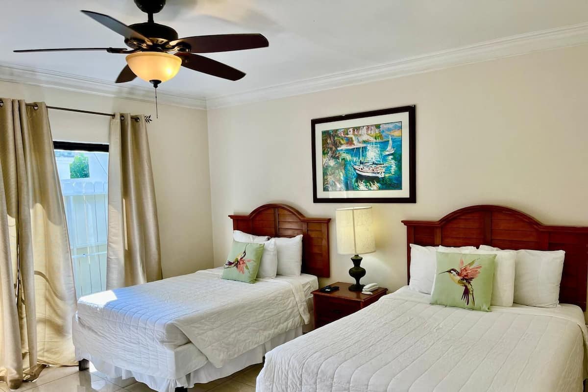 A hotel room with two beds, a ceiling fan, a painting of a coastal scene, and a window with curtains.
