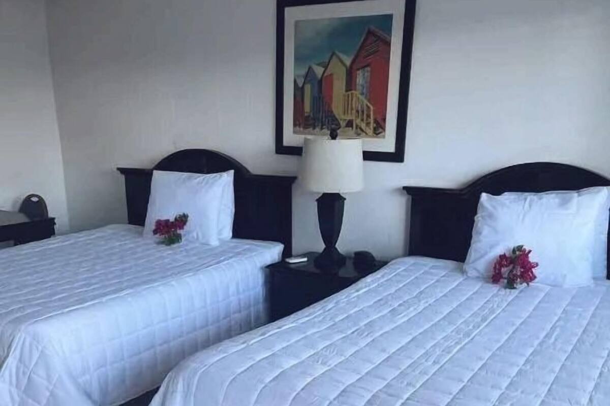 A hotel room with two single beds, a nightstand with a lamp, a framed painting on the wall, and a small table with a chair.