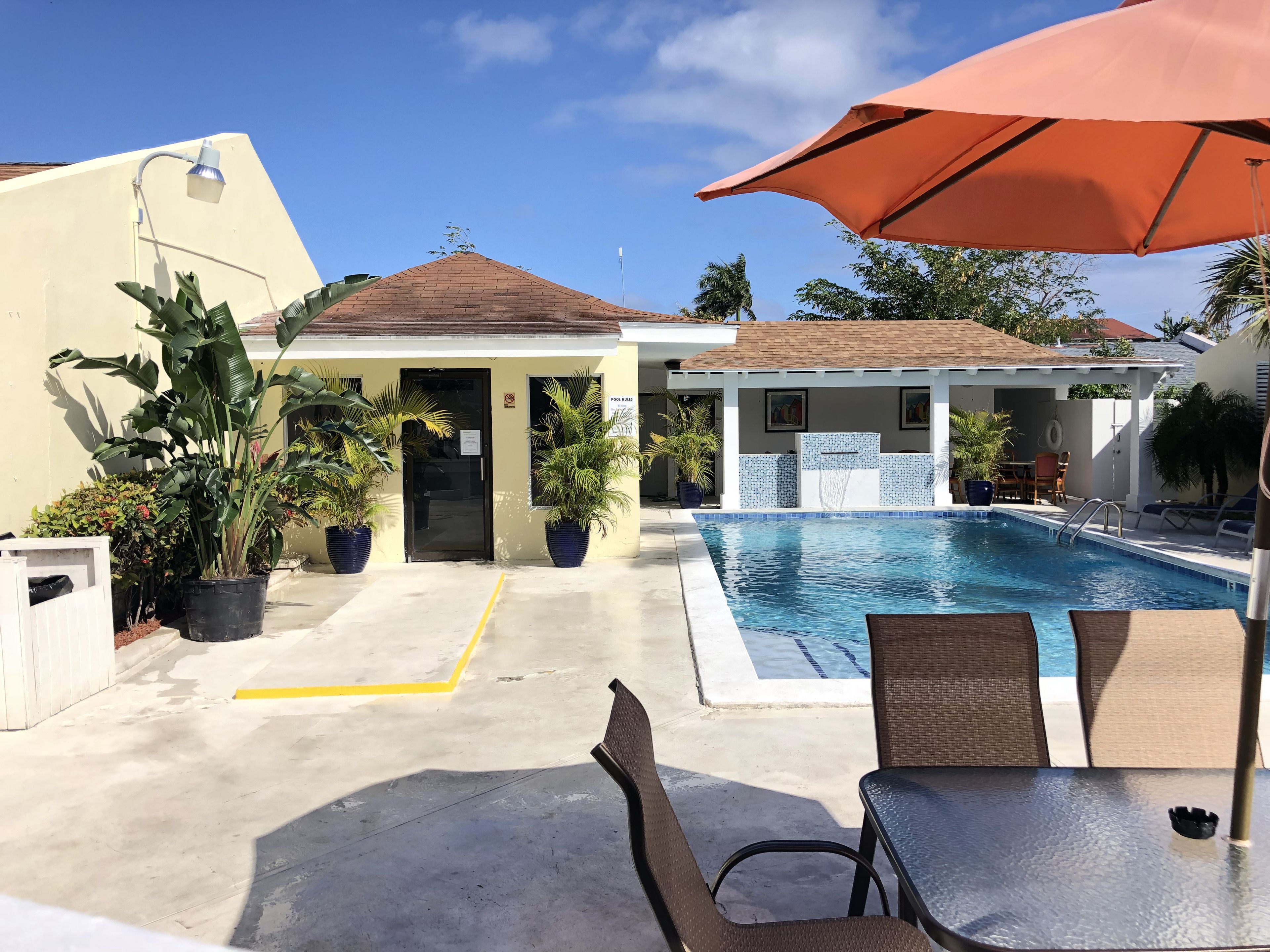 Uma área de piscina com espreguiçadeiras e um guarda-sol, cercada por plantas em vasos e um edifício com fachada em azulejos.