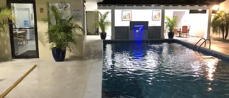 A swimming pool with a blue-lit waterfall, potted plants, and a poolside area with chairs.