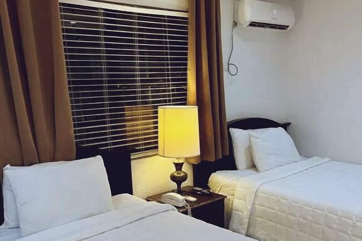 A hotel room with two beds, a window with blinds, a bedside lamp, and an air conditioning unit on the wall.