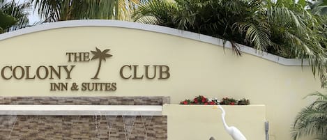 The exterior of The Colony Club Inn & Suites with a sign, a water feature, and tropical landscaping.