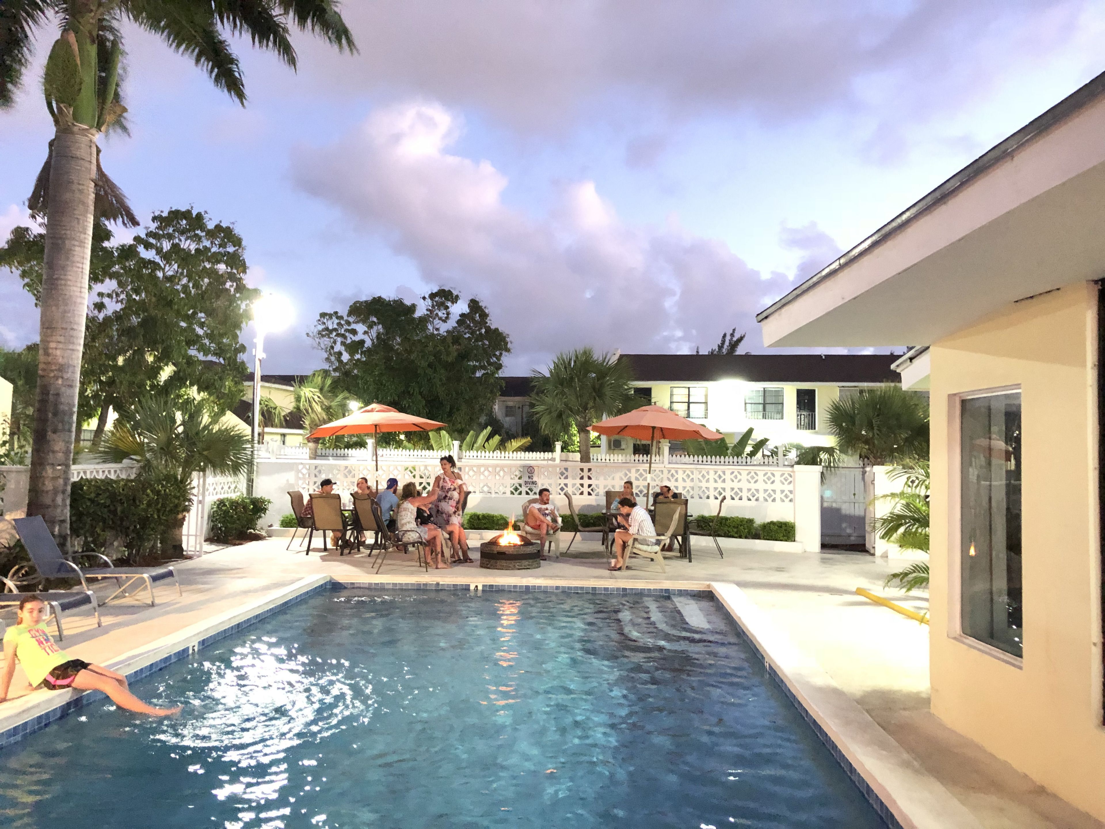A swimming pool area with lounge chairs and umbrellas, surrounded by palm trees and a white fence.
