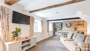 A living room with a sofa, a TV mounted on the wall, a wooden ceiling beam, and a kitchen in the background. - The Pheasant at Neenton (Bridgnorth)