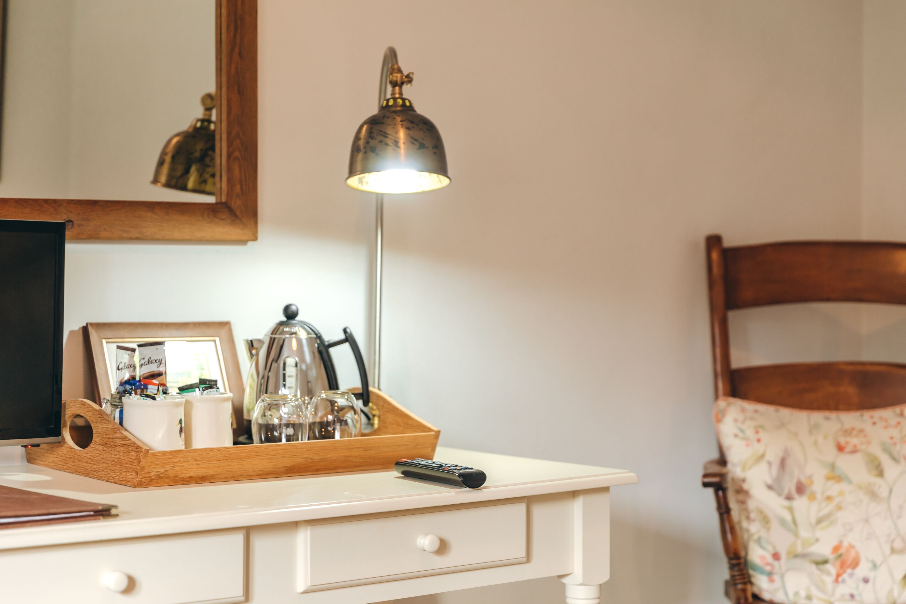 A white desk with a wooden tray holding a remote control, a picture frame, and a kettle. A mirror reflects part of the room.