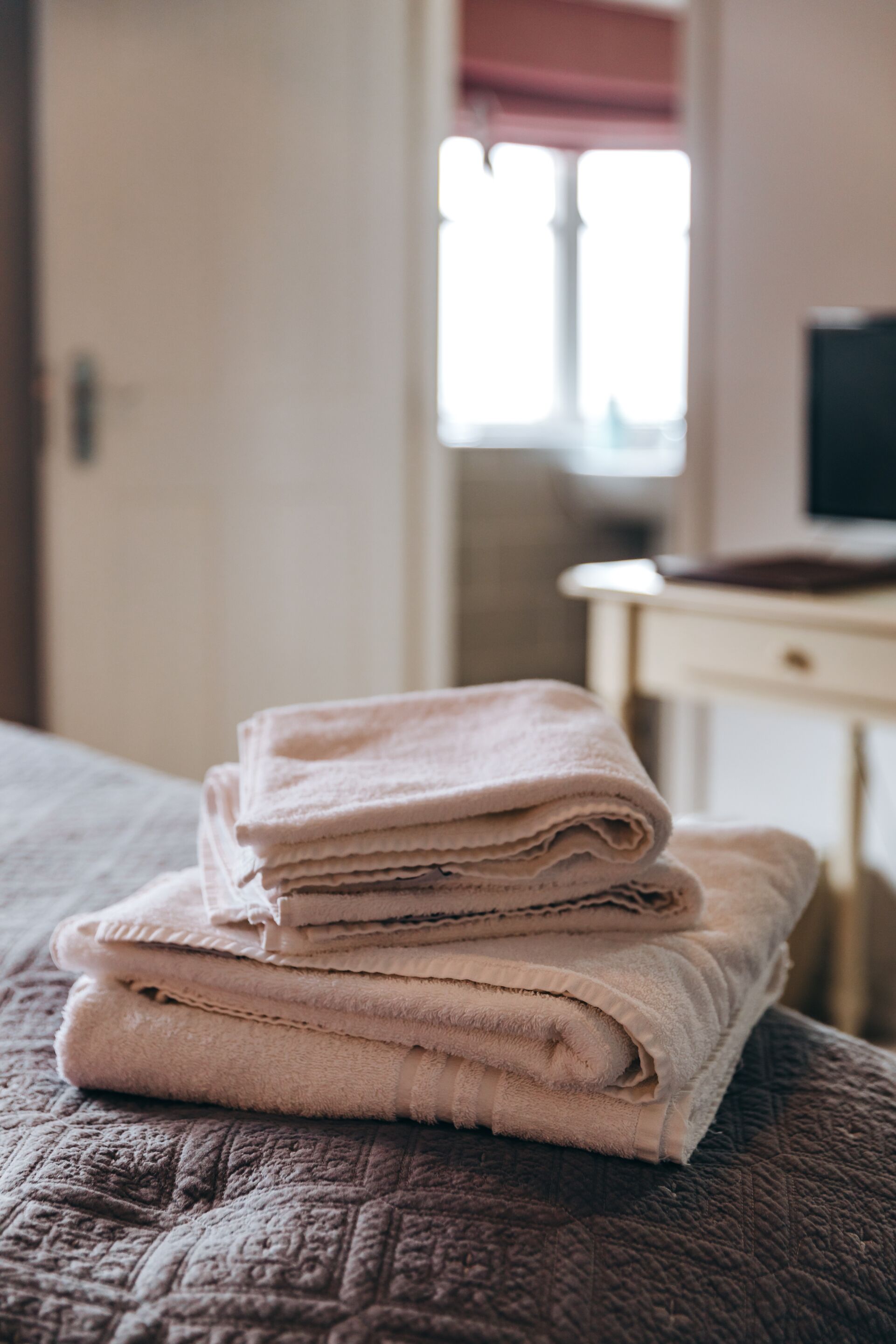 Une pile de serviettes beiges, soigneusement pliées, repose sur un couvre-lit texturé dans une pièce dotée d’une fenêtre et d’une petite commode blanche.