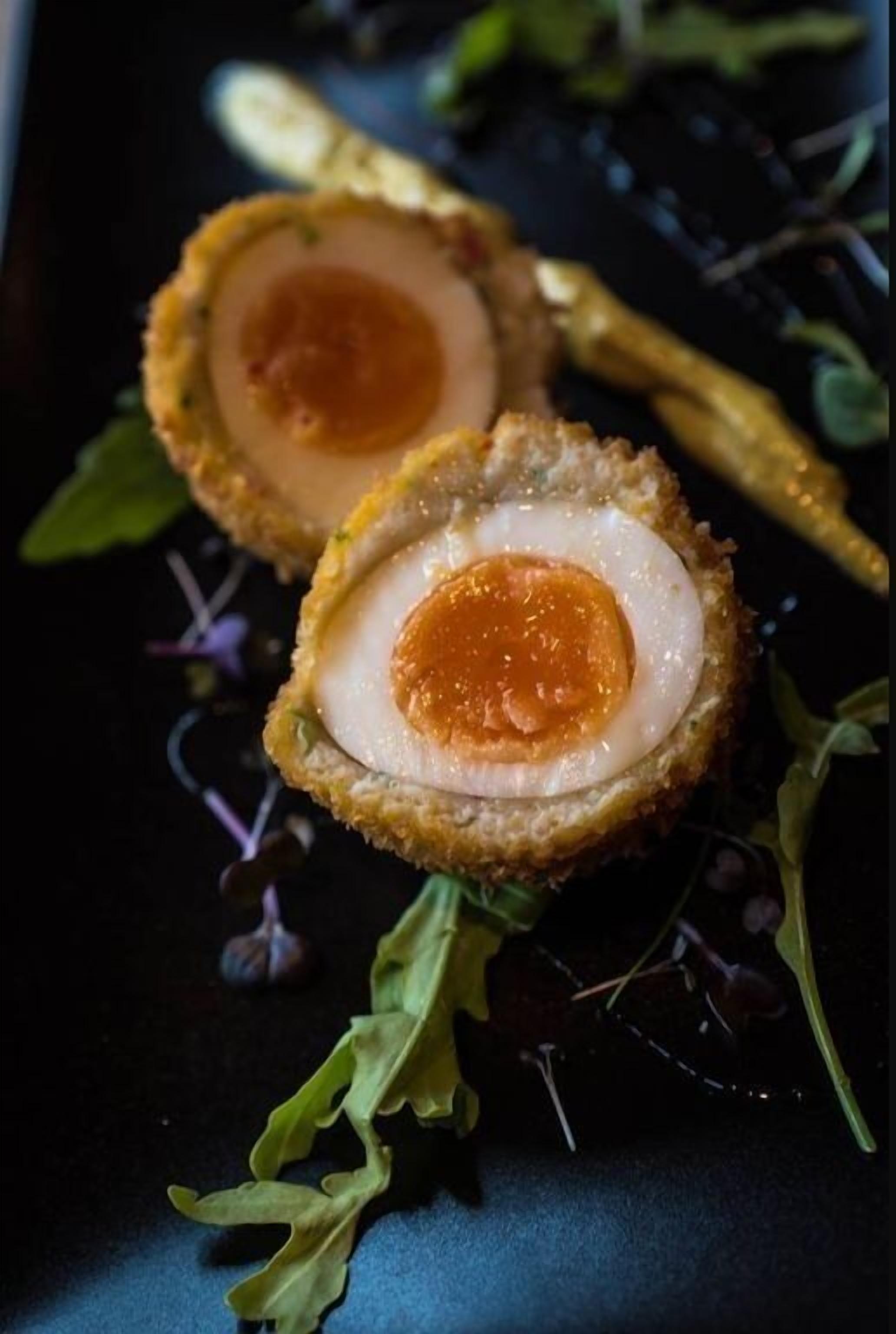 A dish of Scotch eggs with a crispy breadcrumb coating and a soft boiled egg inside, served on a dark plate with some greens.
