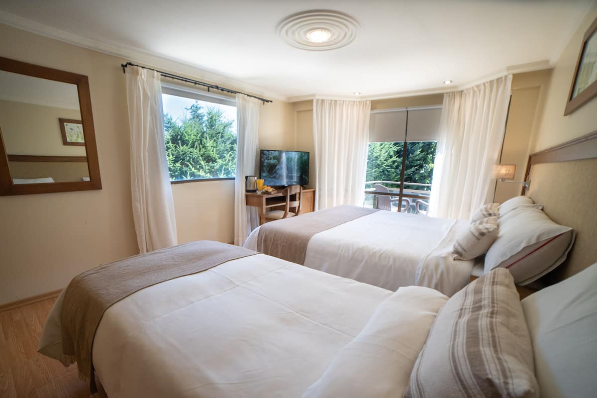 A hotel room with two beds, a TV, a desk with a chair, and a balcony with a view of trees.