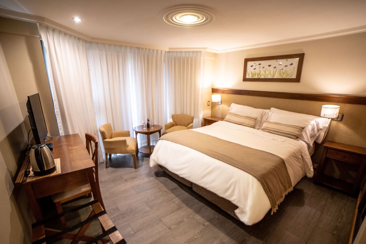 A hotel room with a large bed, a desk with a computer, a chair, and a small table with a lamp.
