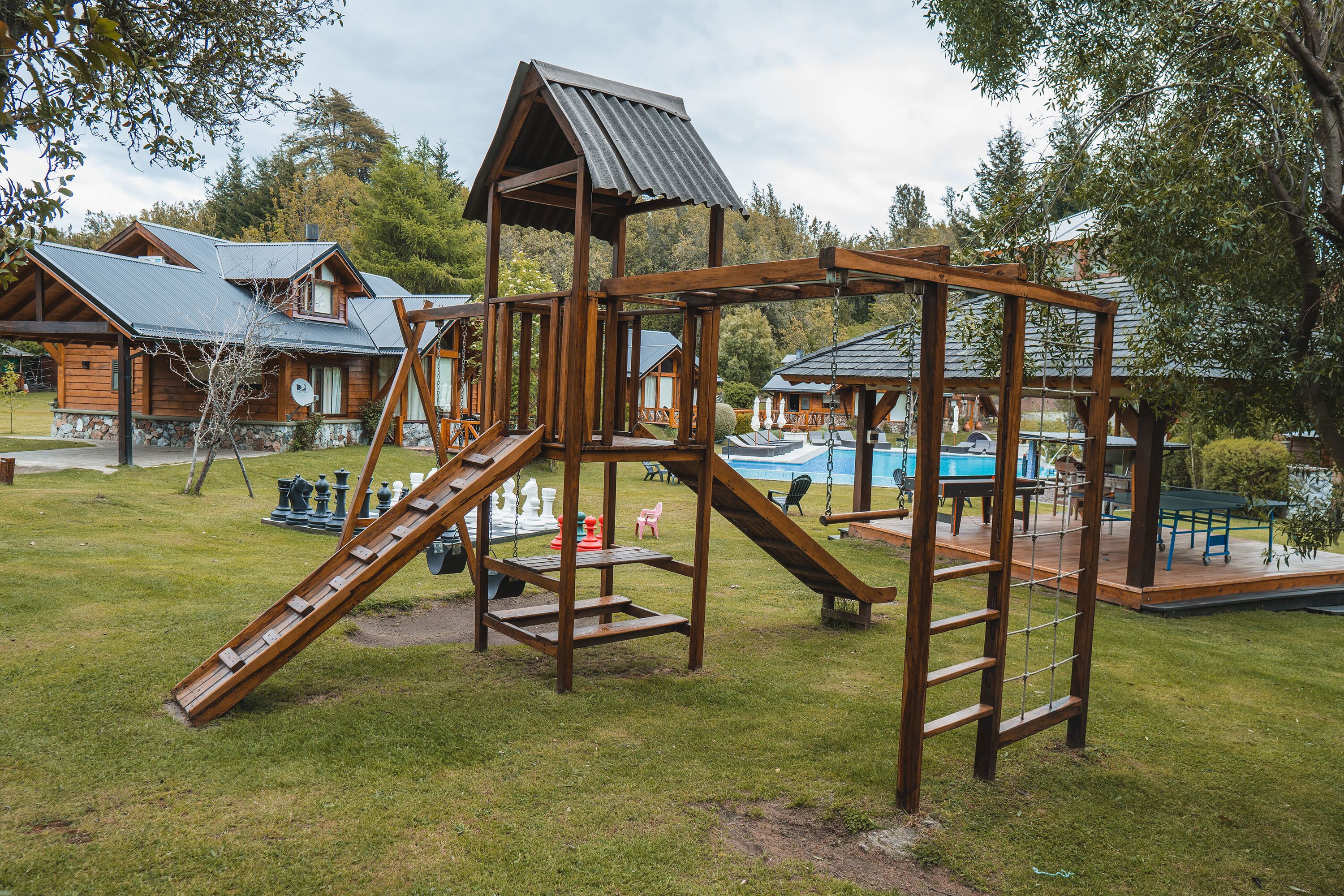 Uma estrutura de madeira para brincar com escorrega e escada de escalada em uma área gramada, com casas ao fundo.