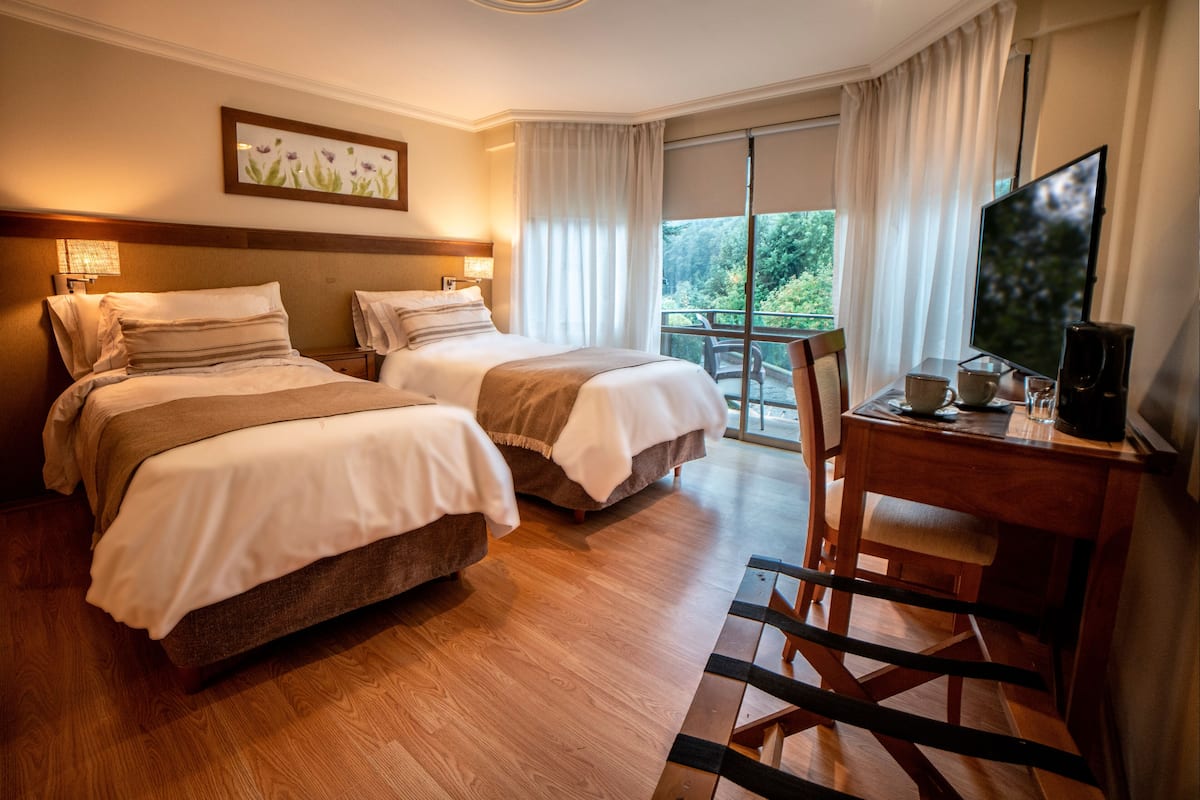 A hotel room with two beds, a desk with a television, and a balcony with a view of greenery.