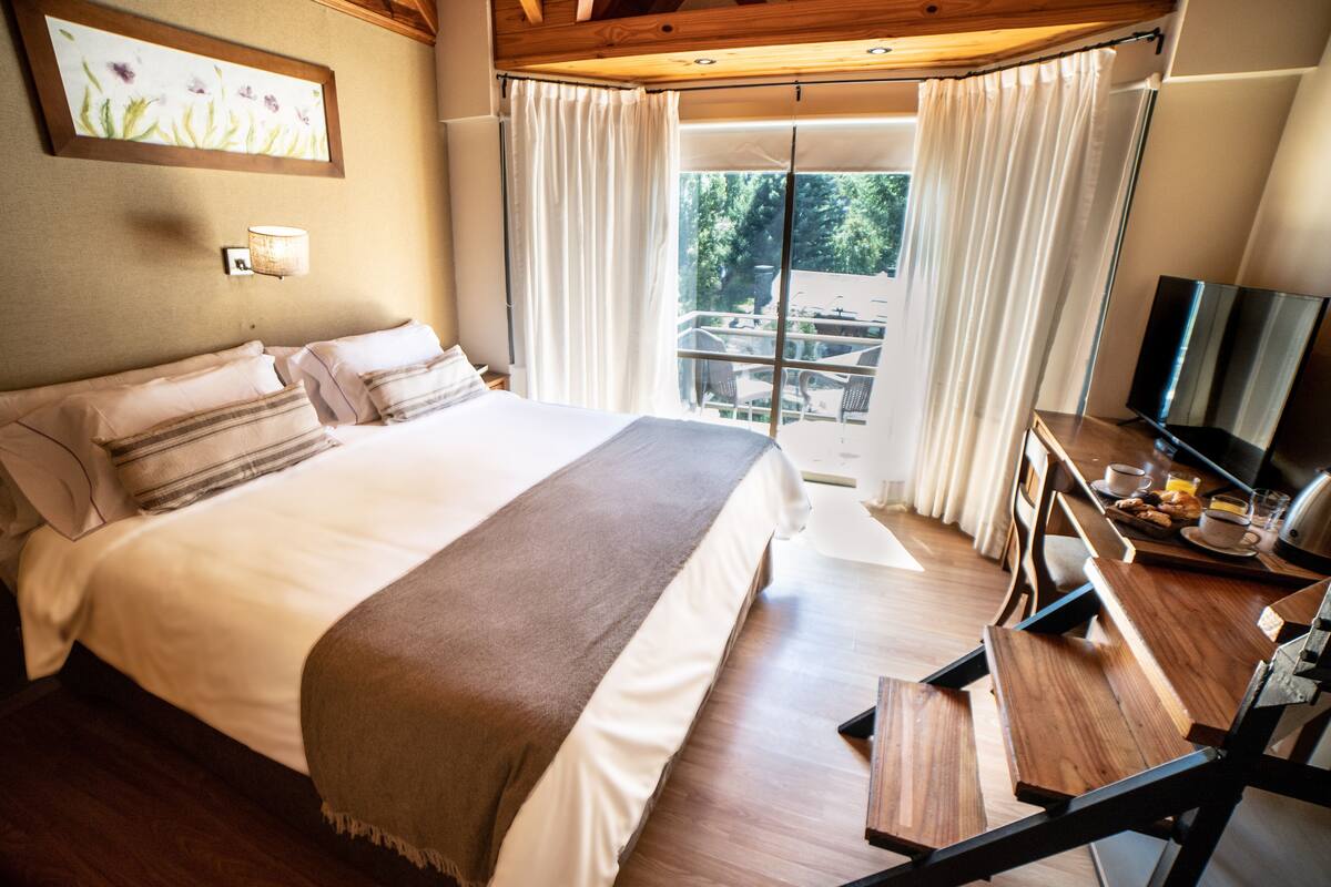 A hotel room with a large bed, a desk with a television, and a balcony with a view of the outdoors.