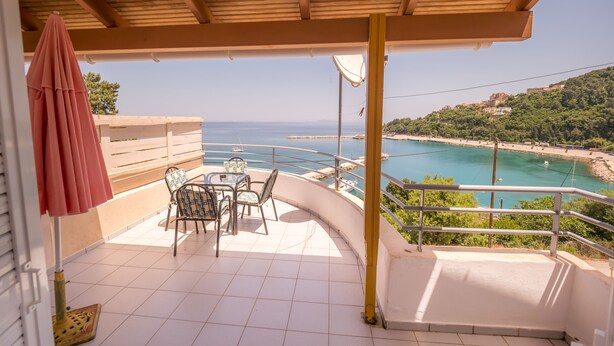 A balcony with a table and chairs, overlooking a coastal area with a clear view of the sea and a beach. - Harbour View - Oceanis Apartments (Kefalonia)
