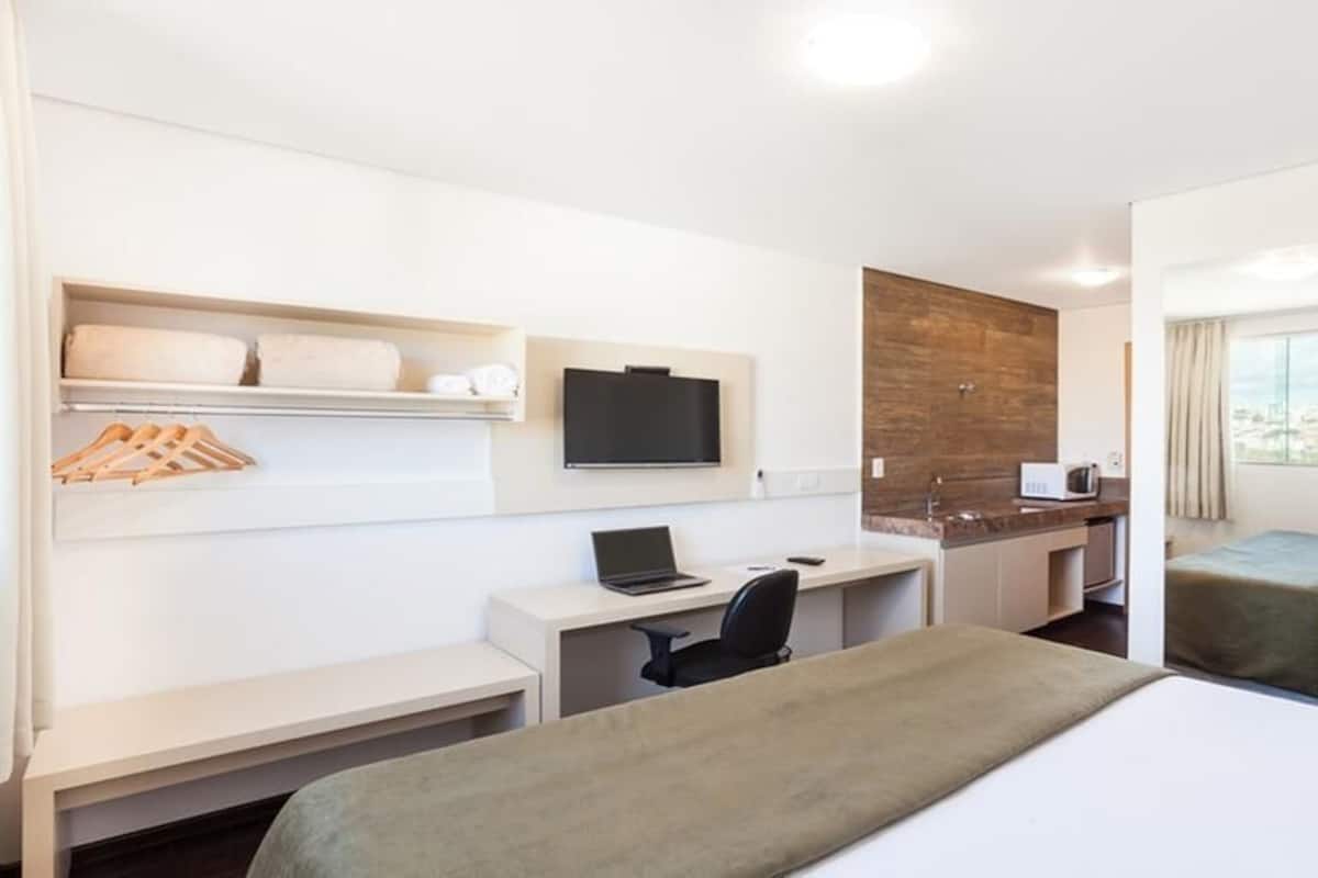 A hotel room with a bed, desk, chair, TV, and a small kitchenette.