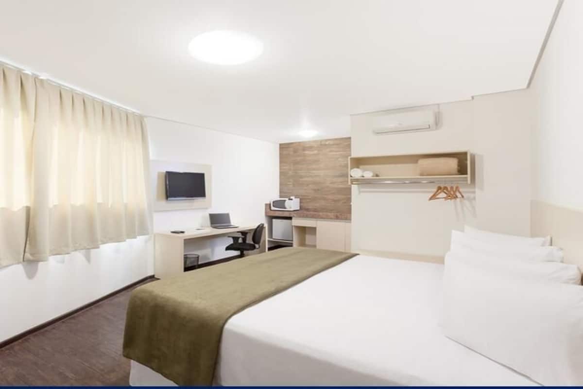 A hotel room with a bed, desk, chair, TV, and a small kitchenette.