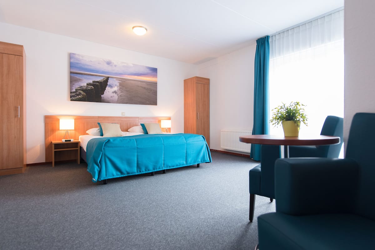 A hotel room with a bed, a desk, a chair, a potted plant, and a painting on the wall.