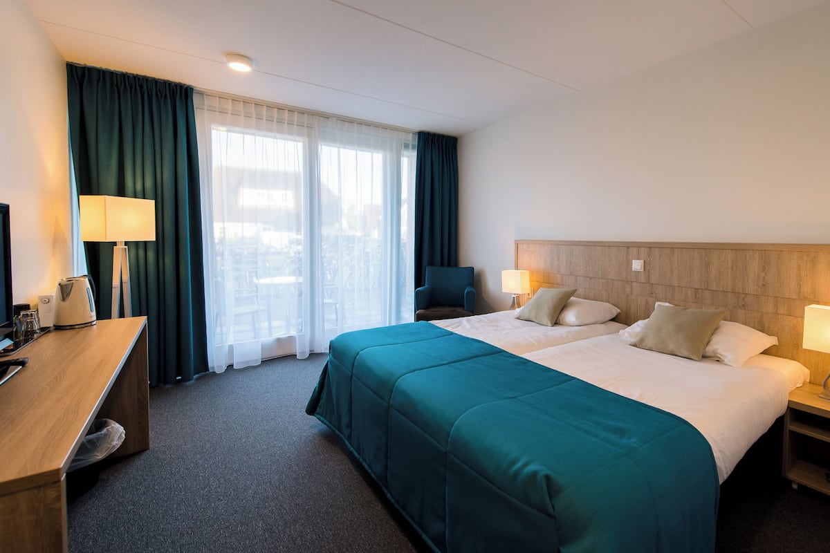 A hotel room with a large bed, two bedside tables with lamps, a desk with a lamp, and a view of the city through the window.